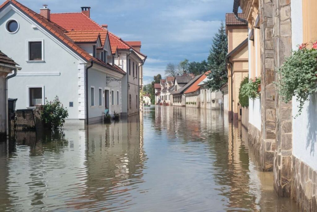 Maisons aux toits rouges et murs blancs partiellement inondées dans une rue, illustrant la préparation des communes aux inondations en Belgique.