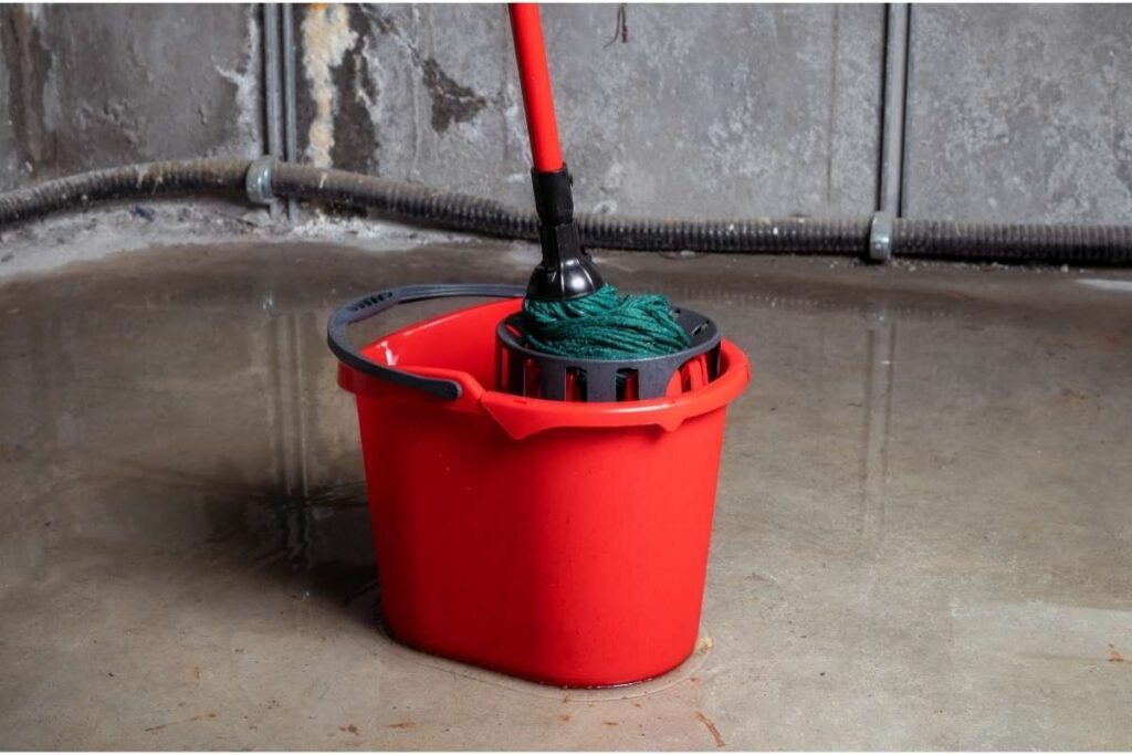 Un seau rouge avec une serpillière verte est posé sur le sol en béton d'une cave humide, illustrant la protection des caves contre les inondations.